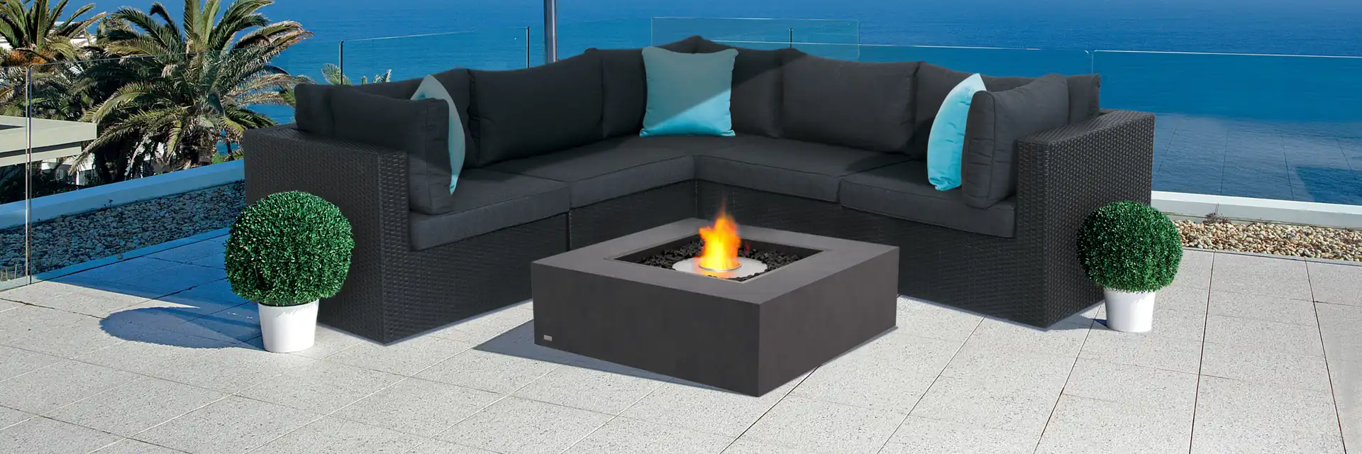 EcoSmart Base 40 fire table burning in front of a dark corner sofa on a pale terrace, under a blue parasol with the sea behind
