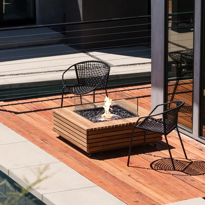 EcoSmart Base 40 fire table in teak burning on a timber deck beside a dark modern house, with two wire chairs