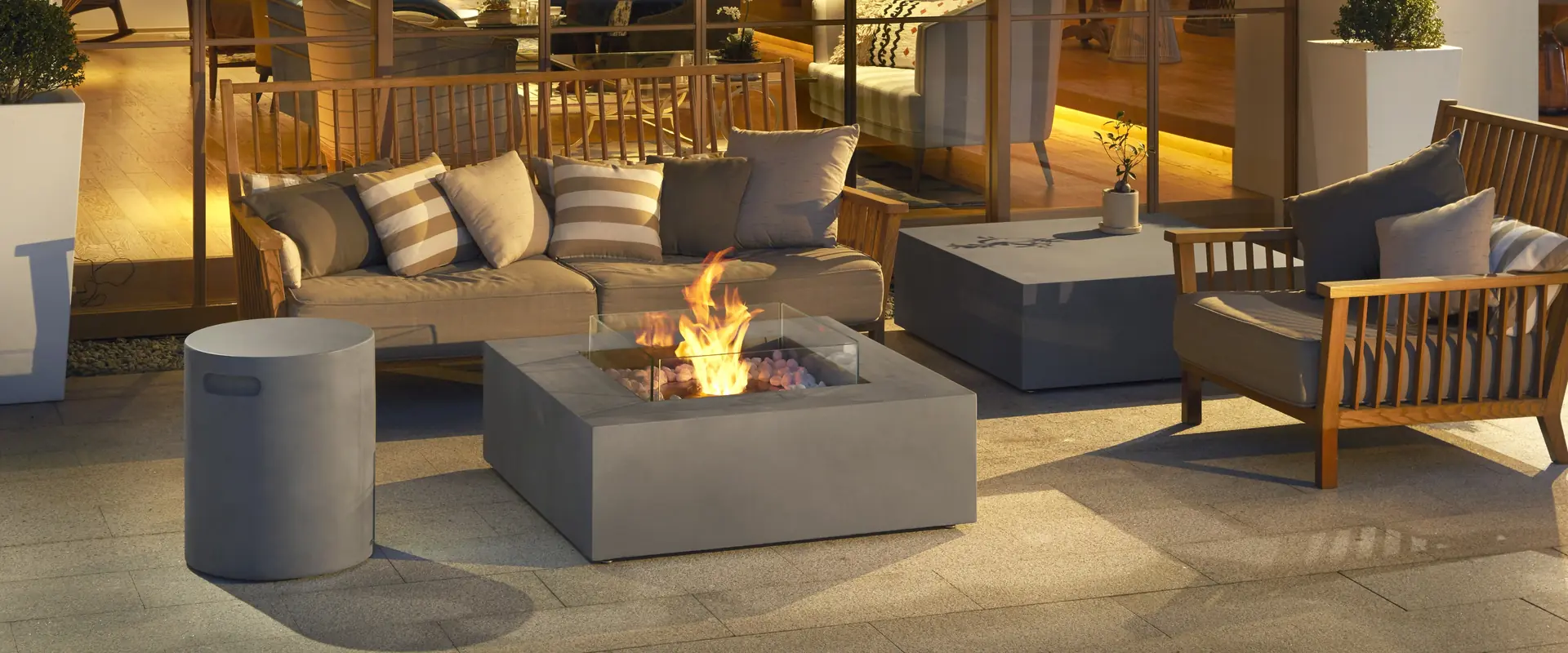 Square concrete EcoSmart Base bioethanol fire table with a glass screen in the middle of a terrace lounge with striped cushions, in front of a lit glass-walled lounge in the evening