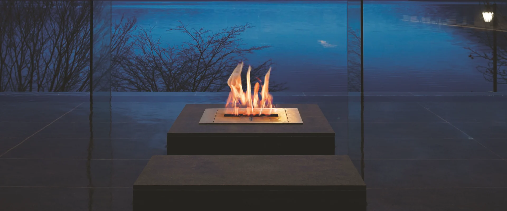 Flames rising from an EcoSmart bioethanol burner set in a dark stone block, in front of floor-to-ceiling glass with a view of a lake and hills at dusk