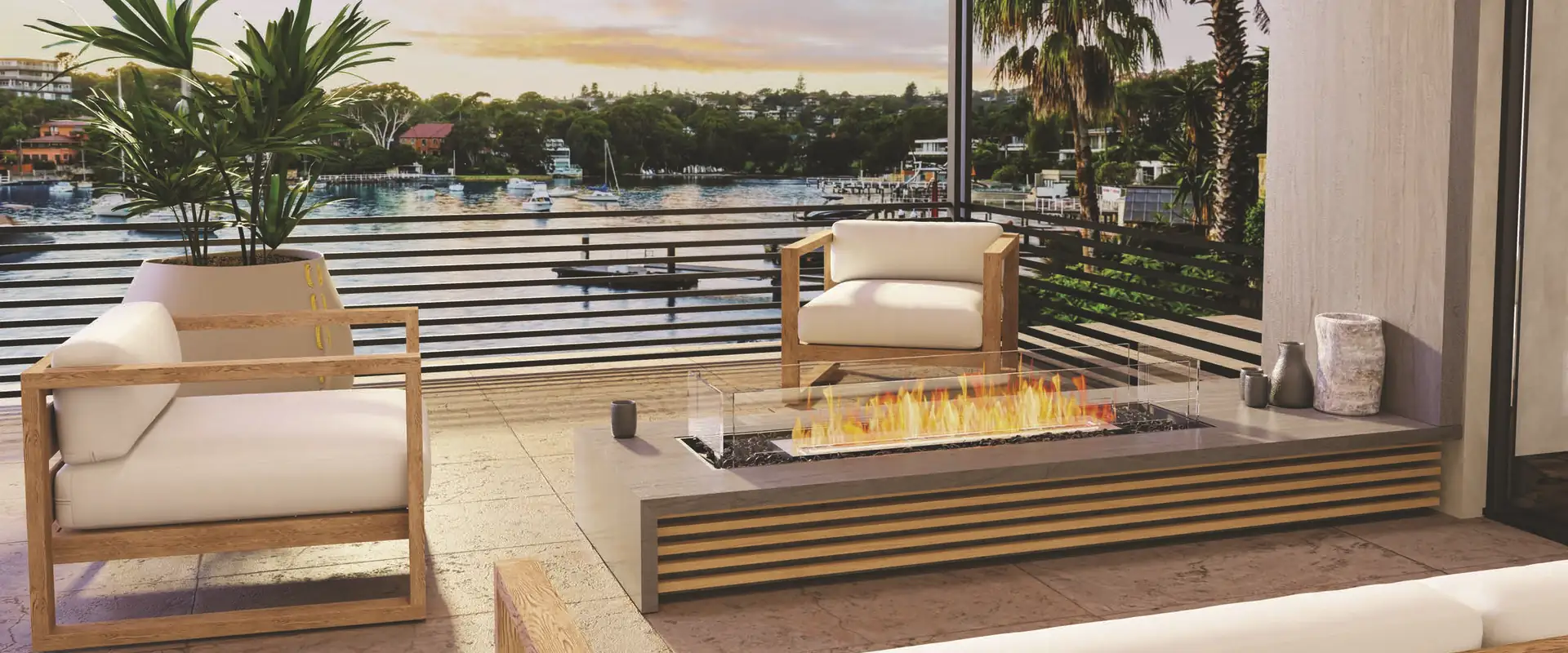 EcoSmart bioethanol fire with a long flame behind a glass screen, built into a low concrete and timber table on a covered terrace with white armchairs, overlooking a harbour with palm trees at sunset