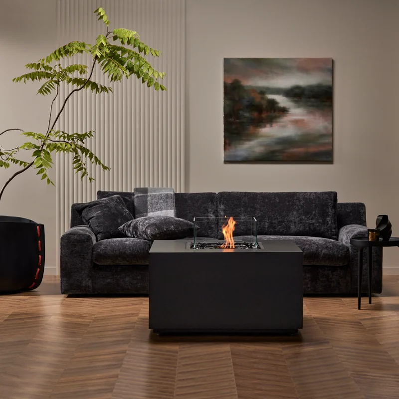 EcoSmart Chaser 38 fire table in graphite burning in front of a dark velvet sofa, beside a tall plant