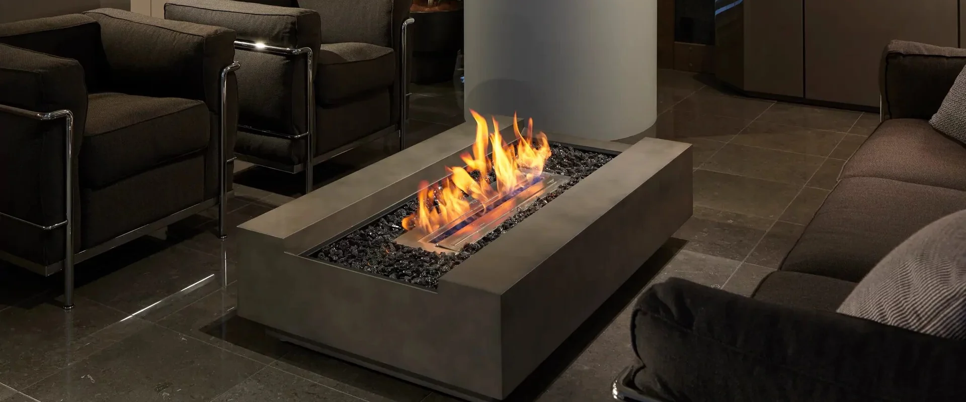 Rectangular concrete EcoSmart Cosmo 50 bioethanol fire table with a long flame in a dark-toned living room, between black leather armchairs and a sofa