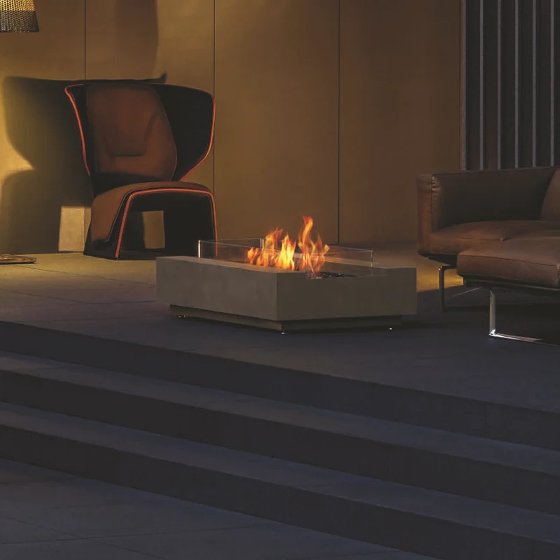Rectangular concrete EcoSmart Cosmo 50 bioethanol fire table with a glass screen burning on a raised terrace at night, beside a leather armchair and a woven floor lamp