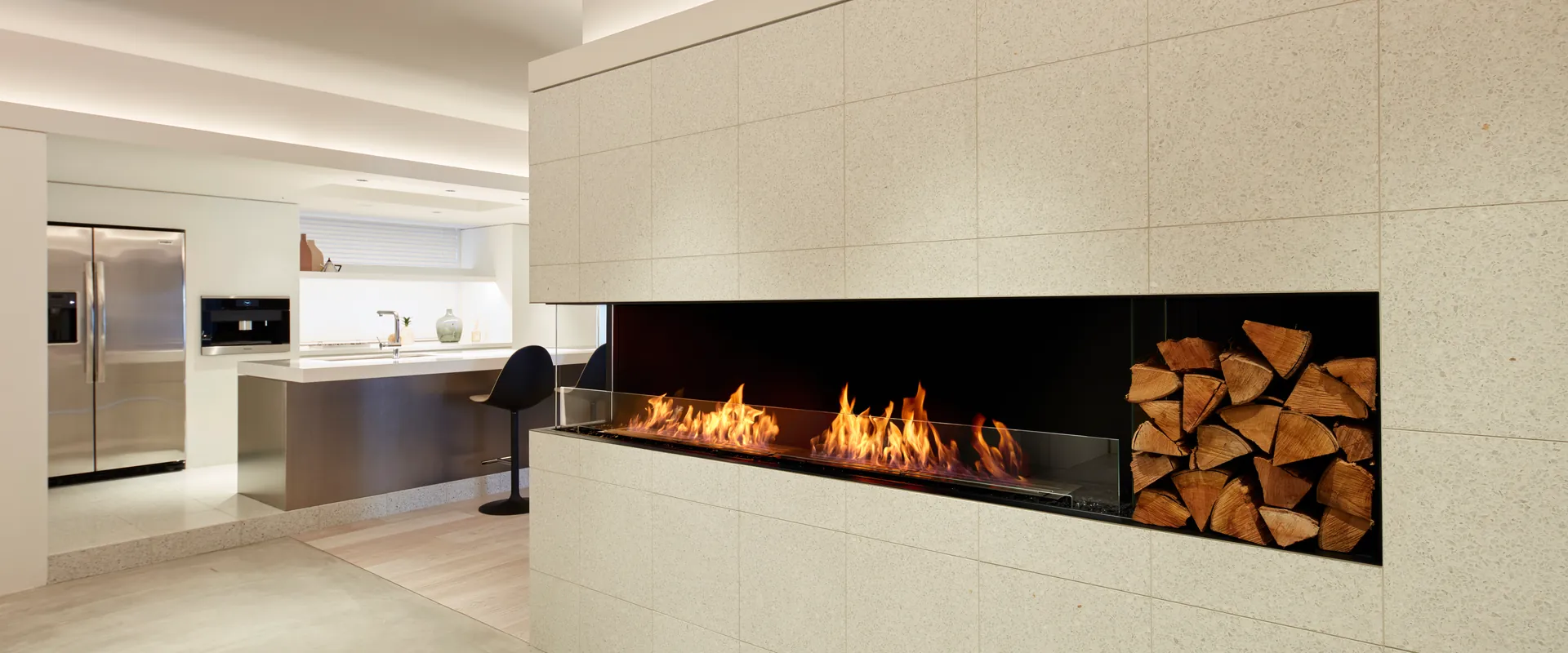 EcoSmart Flex bioethanol corner fireplace with a log store set into a pale stone wall, in an open-plan living space with a kitchen behind