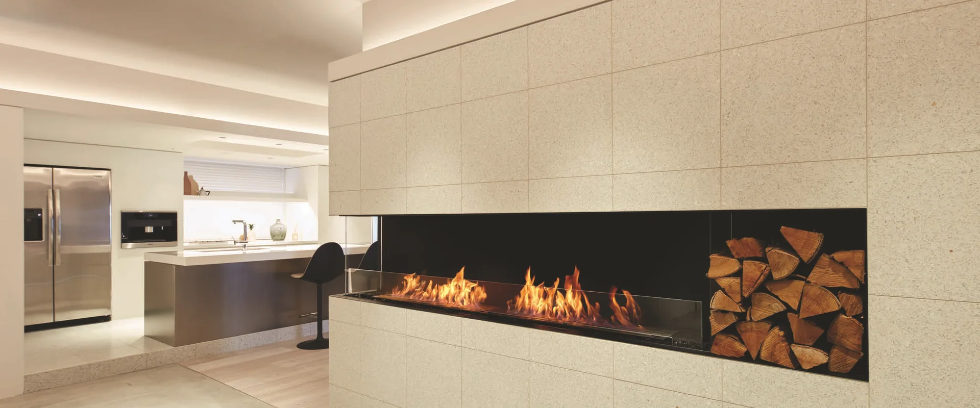 Long EcoSmart Fire Flex bioethanol corner fireplace with a stacked-log niche, built into a pale stone-clad wall beside an open kitchen