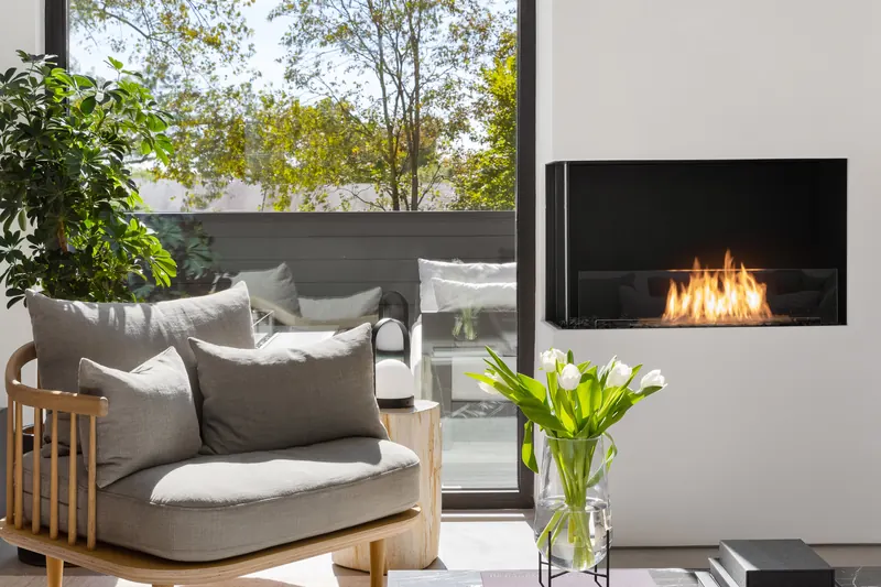 EcoSmart Fire Flex corner fireplace burning in a white wall beside a tall window with trees, behind a gray armchair