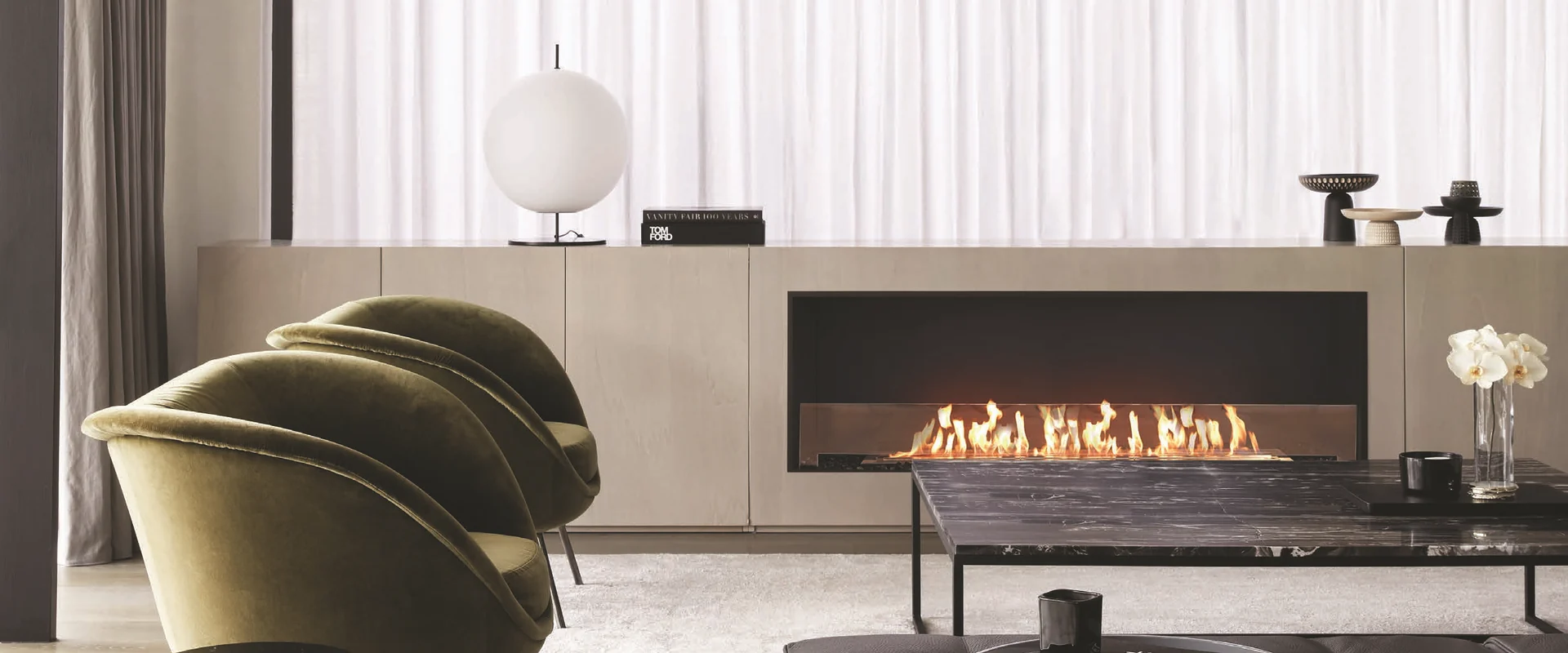 Long EcoSmart Fire Flex bioethanol fireplace built into a pale stone wall unit in a bright living room, with two green velvet armchairs and a marble coffee table in front