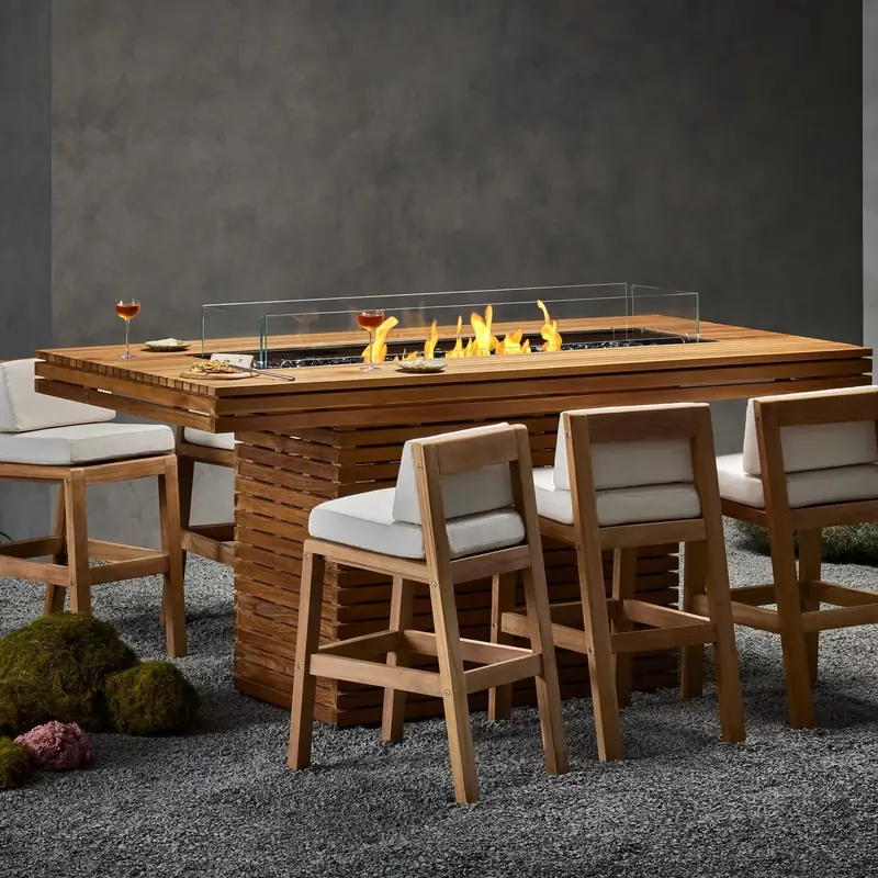 Teak EcoSmart Gin 90 Bar bioethanol fire table with a long flame behind a glass screen, surrounded by teak bar stools with white cushions on a gravel garden with moss mounds