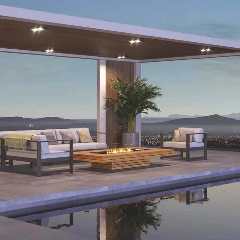 Teak EcoSmart Gin 90 Low bioethanol fire table between a sofa and armchairs under a white pergola, beside a reflecting pool with mountains beyond at dusk