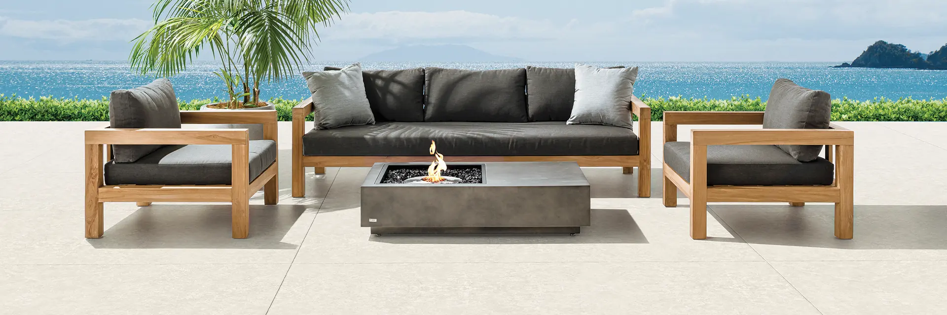 EcoSmart Manhattan 50 fire table burning between teak sofas on a pale deck by the beach, with palms and the sea behind