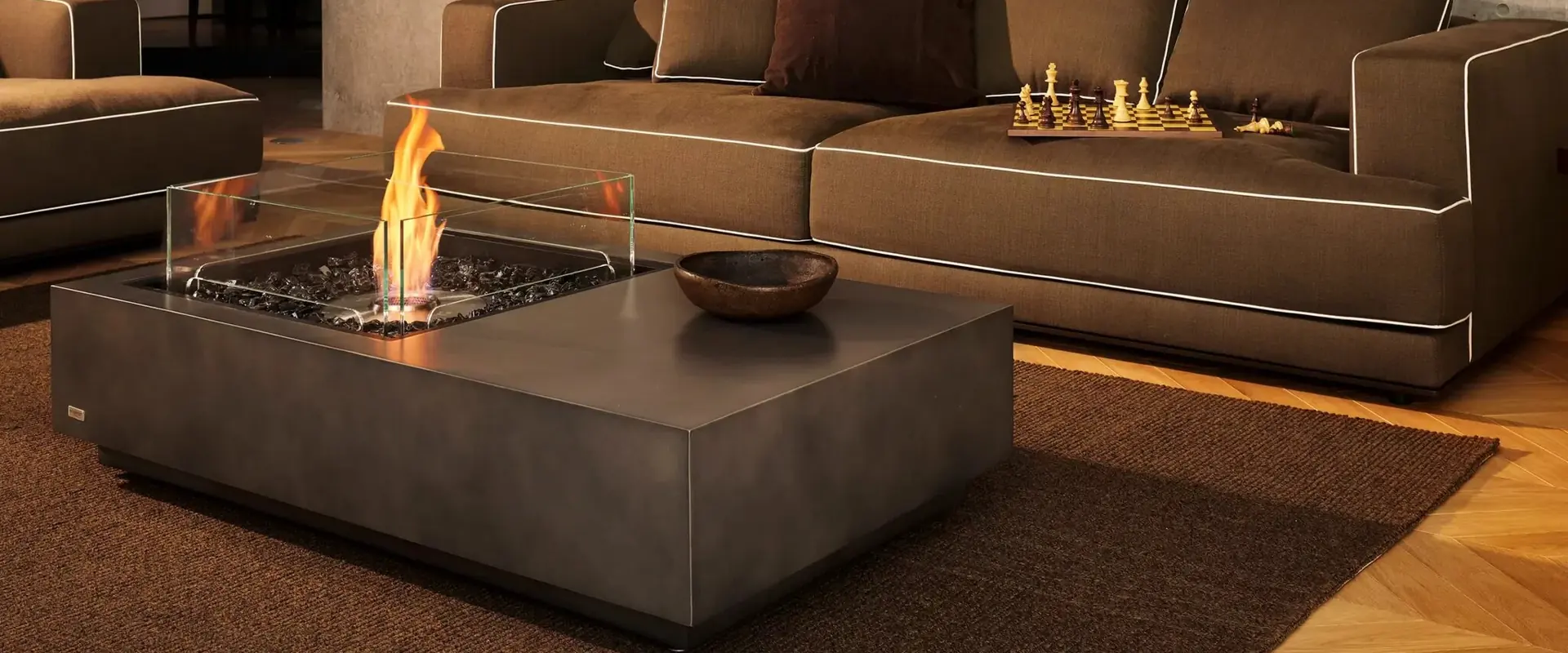 Square concrete EcoSmart Manhattan 50 bioethanol fire table with a glass screen burning on a rug in front of a brown sofa, against a tall board-formed concrete wall