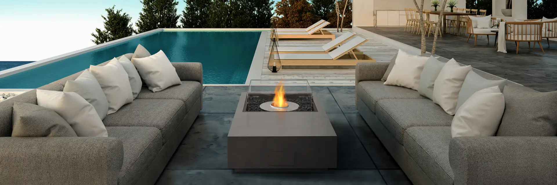 EcoSmart Manhattan 50 fire table burning between gray sofas on a terrace beside an infinity pool, with pine trees and the sea beyond