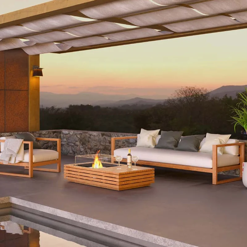 Teak EcoSmart Manhattan 50 bioethanol fire table with a glass screen between a sofa and an armchair on a pergola-covered terrace, with hills beyond at sunset