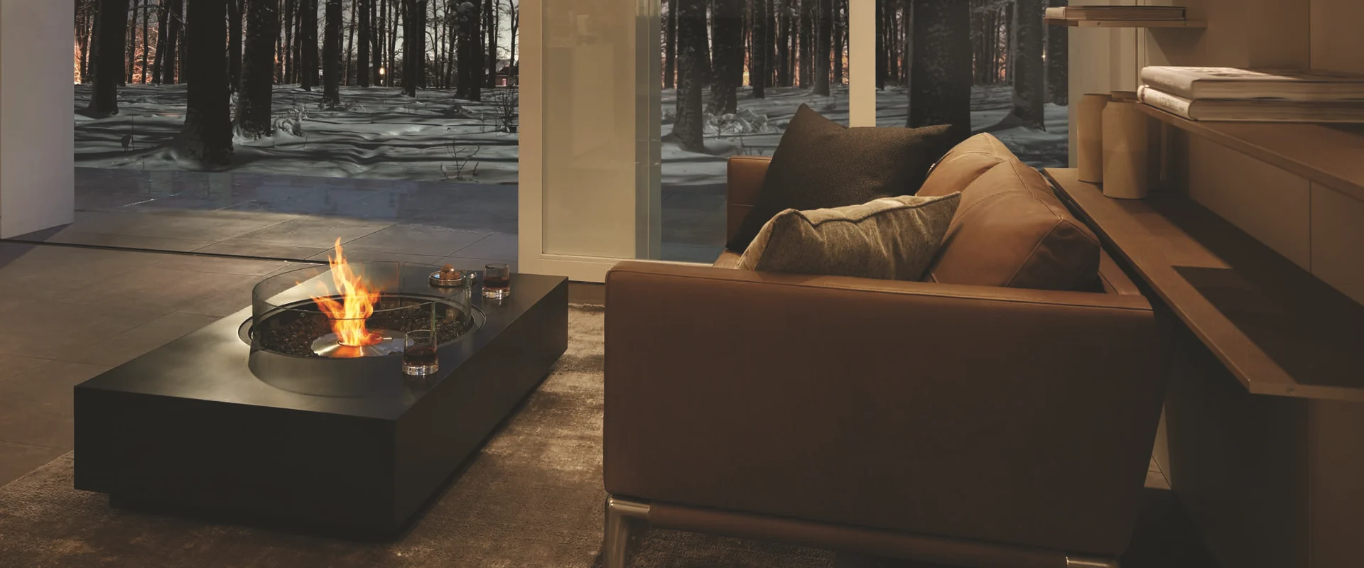 Black EcoSmart Martini 50 bioethanol fire table with a round glass screen burning in a living room beside a leather sofa, with tall windows onto a snowy forest