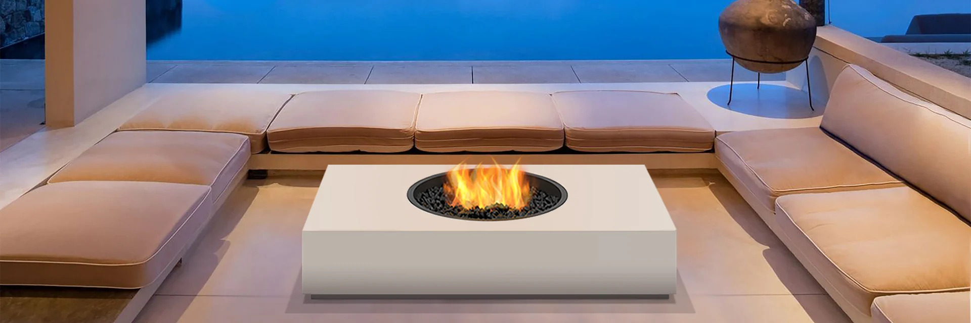 EcoSmart Martini 50 fire table in white burning in a sunken lounge with cushioned seating, open to an infinity pool and a bay at dusk