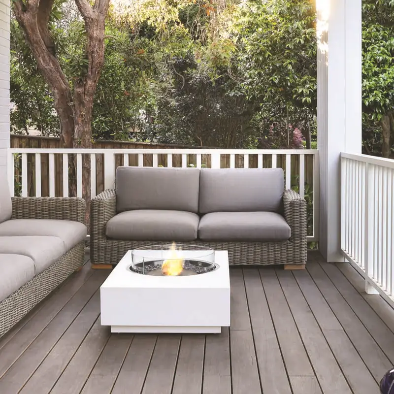 White EcoSmart Martini 50 bioethanol fire table with a round glass screen on the timber deck of a white veranda, in front of wicker sofas and a leafy garden