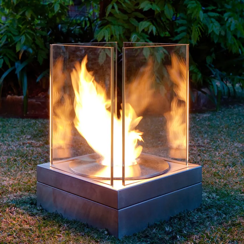 EcoSmart Mini T bioethanol fire with a tall flame inside its glass screens on a stainless steel base, standing on a garden lawn in front of dense foliage at dusk