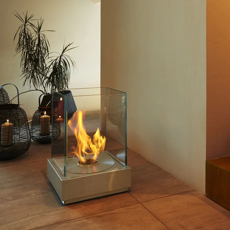 EcoSmart Mini T bioethanol fire burning inside its glass screens on a tiled floor at the foot of a timber staircase, beside wicker lanterns and a potted palm