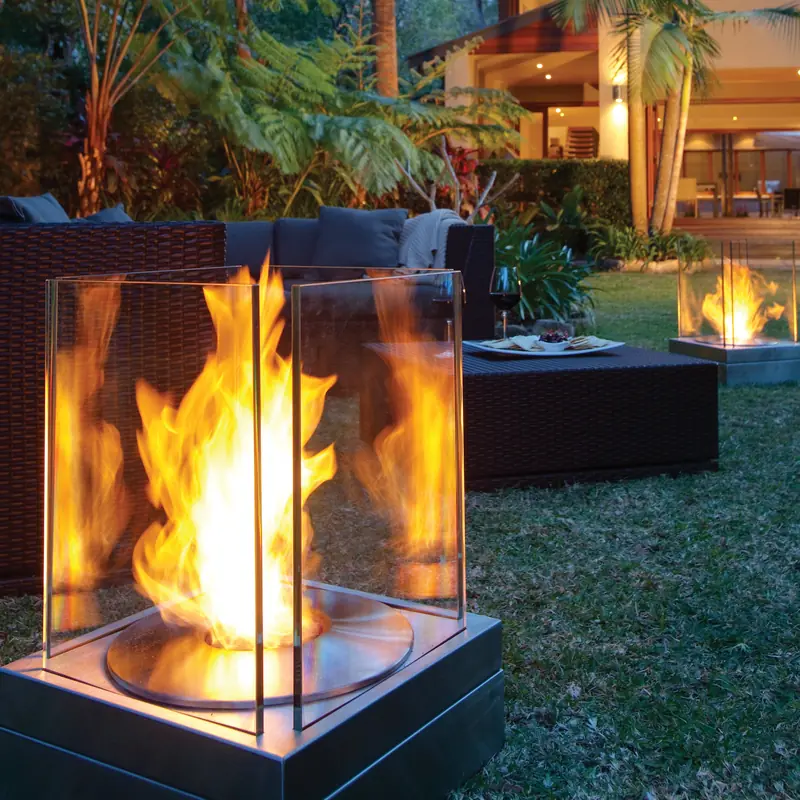 EcoSmart Mini T bioethanol fire with tall flames behind its glass screens on a garden lawn at dusk, with a second one beside an outdoor sofa and a lit house behind
