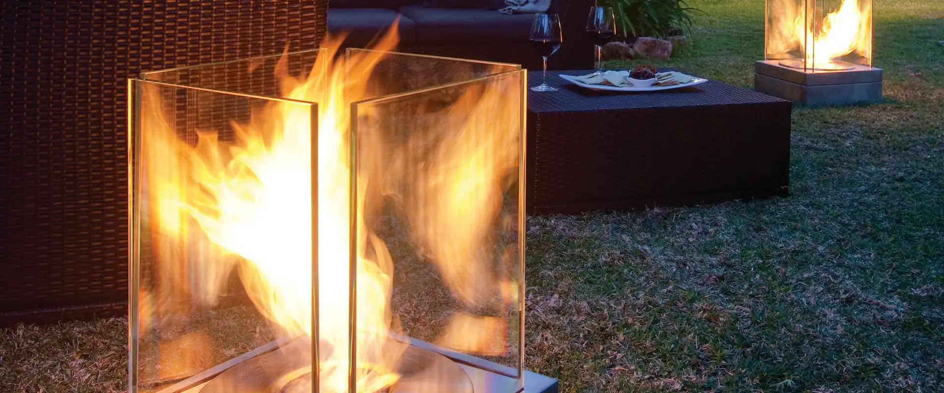 Two EcoSmart Mini T bioethanol fires with flames behind glass screens on a garden lawn at dusk, beside a dark wicker sofa, with a lit house among palms behind