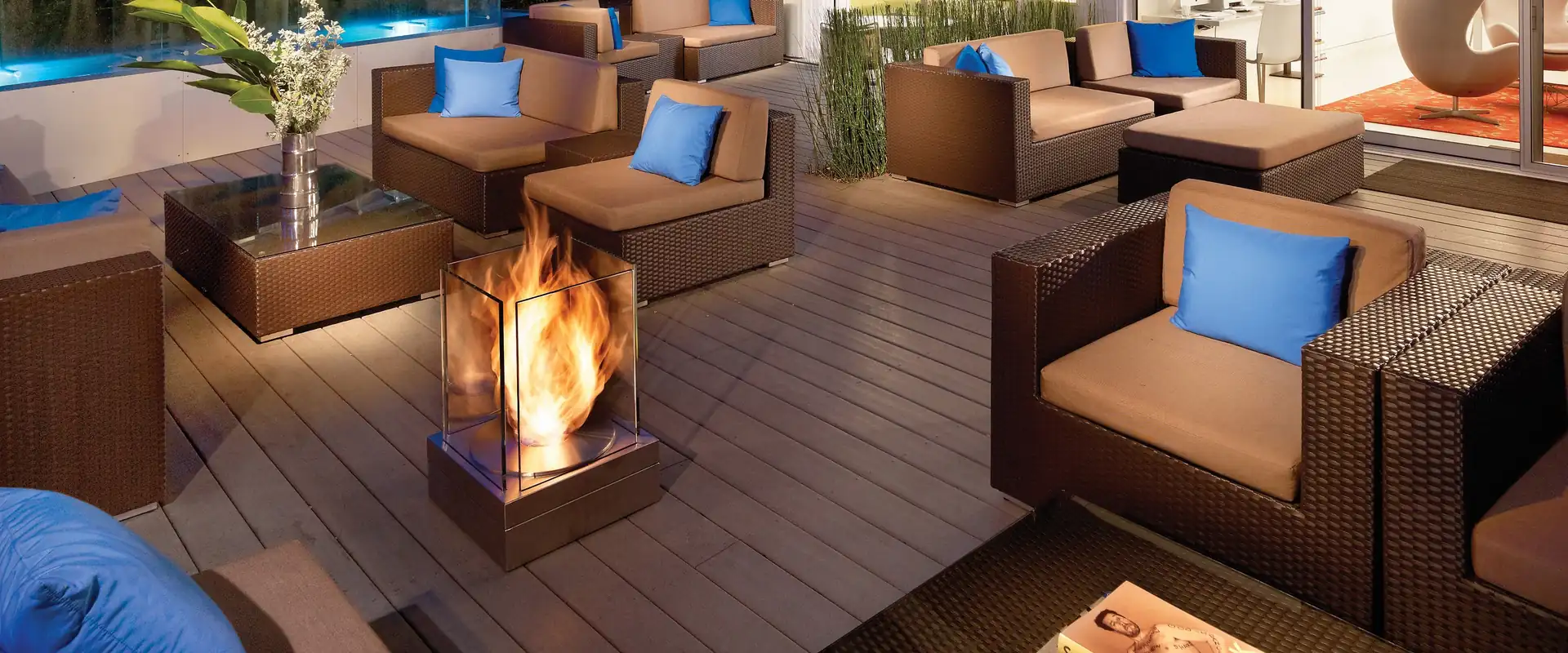 EcoSmart Mini T bioethanol fire glowing among wicker lounge chairs with blue cushions on the timber deck of a modern house, with a pool behind at dusk