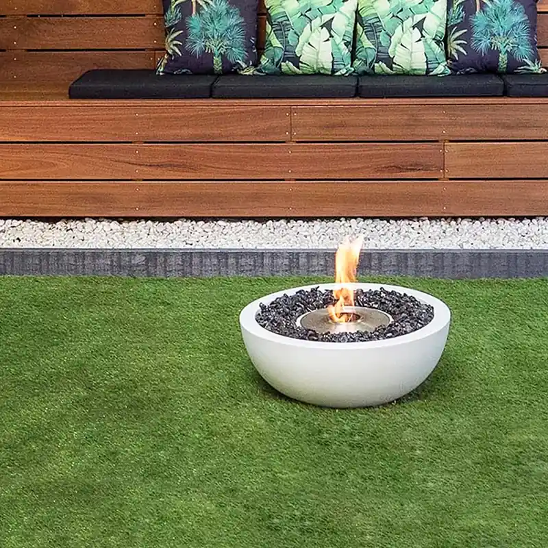 White EcoSmart Mix 600 bioethanol fire bowl burning on a lawn in front of a timber-slat garden wall with a built-in bench and leaf-print cushions