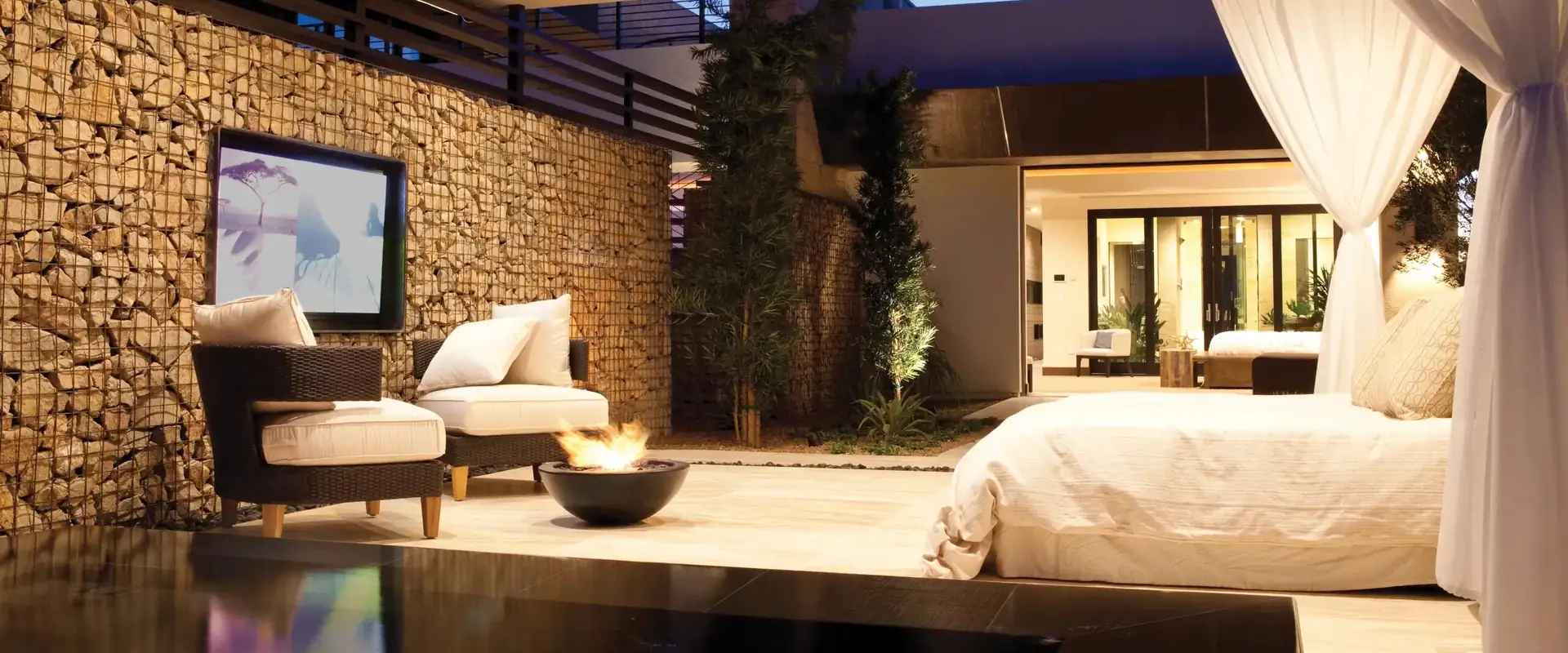 Dark EcoSmart Mix 600 bioethanol fire bowl burning on the stone floor of an open pavilion at night, beside a daybed with white curtains, a lounge chair and a dark pool