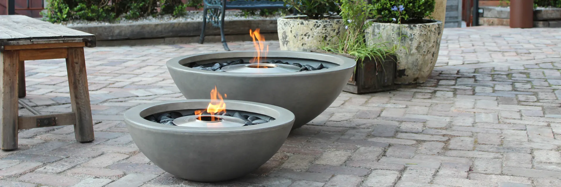 Two EcoSmart Mix fire bowls in concrete gray burning on a cobbled courtyard beside a wooden bench