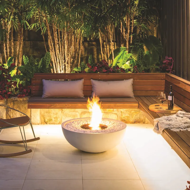 White EcoSmart Mix 850 bioethanol fire bowl glowing on a paved courtyard at night, beside a rocking chair and a timber bench backed by bamboo