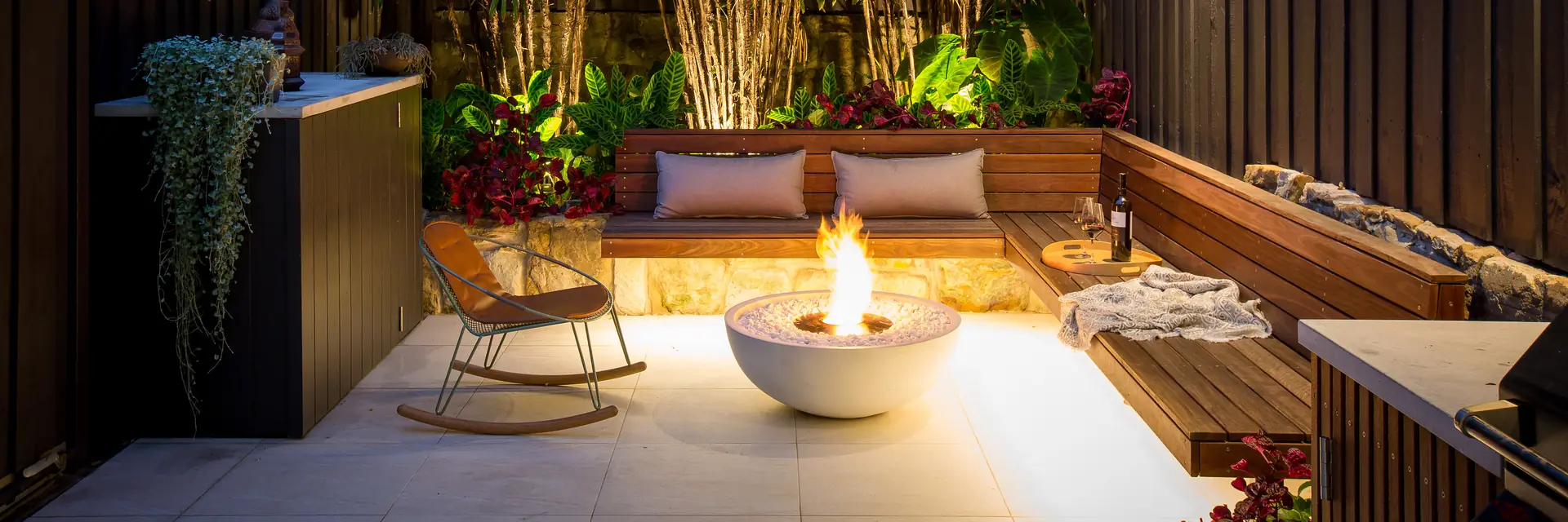 EcoSmart Mix 850 fire bowl in white burning in a courtyard with a timber bench, a rocking chair and bamboo lit at night