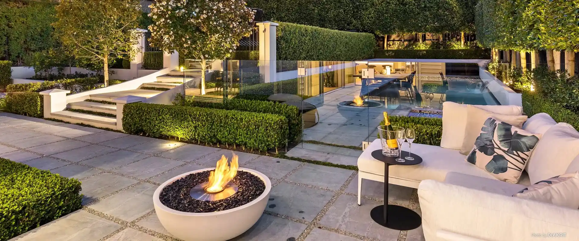 Pale EcoSmart Mix 850 bioethanol fire bowl burning on a paved garden terrace with clipped hedges and a white outdoor sofa, with steps, a pool and a house beyond at dusk