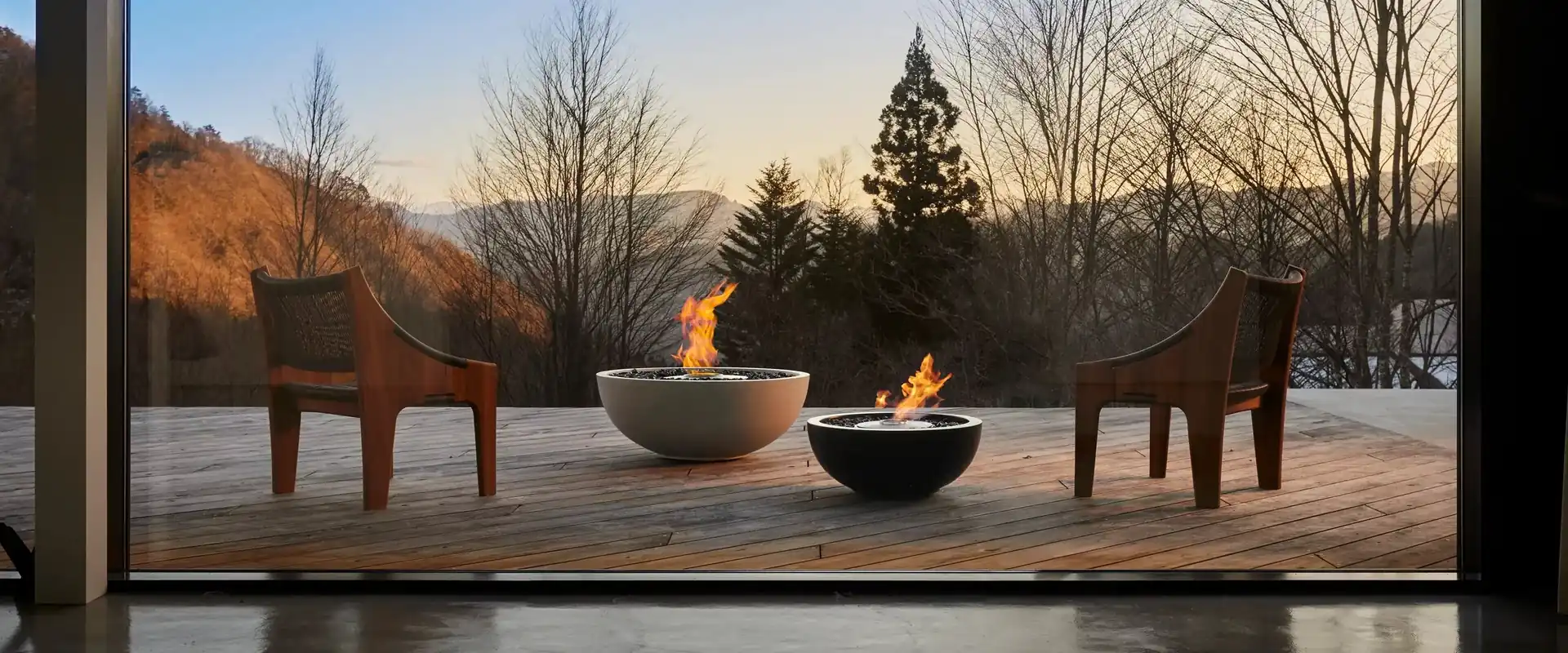 Two EcoSmart Mix bioethanol fire bowls, one pale and one dark, burning between two wooden armchairs on a timber deck overlooking wooded hills at dusk
