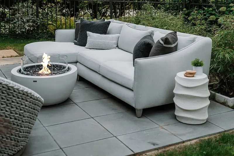 EcoSmart Pod 30 fire pit in white burning beside a pale outdoor sofa on a stone terrace, with greenery behind