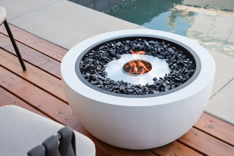 Close view of an EcoSmart Pod 30 fire pit in white with black glass, burning on a timber deck beside a pool