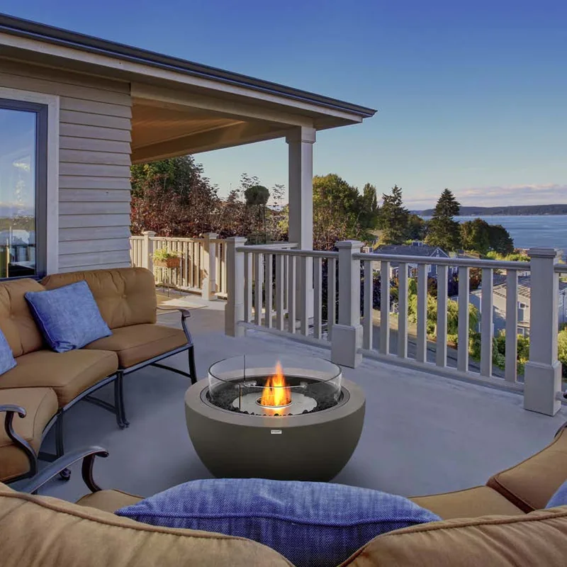 Dark EcoSmart Pod 30 bioethanol fire pit with a glass screen on the veranda of a house, between cushioned sofas, overlooking a bay at dusk