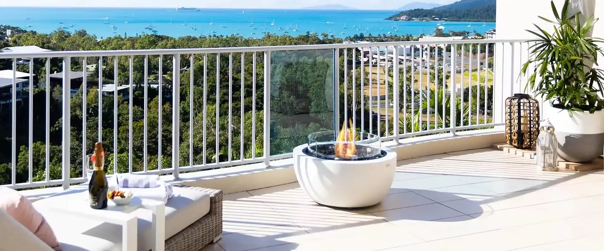 White EcoSmart Pod 30 bioethanol fire pit with a glass screen on a sunny white-tiled balcony with a wicker daybed, overlooking a green hillside and a turquoise bay