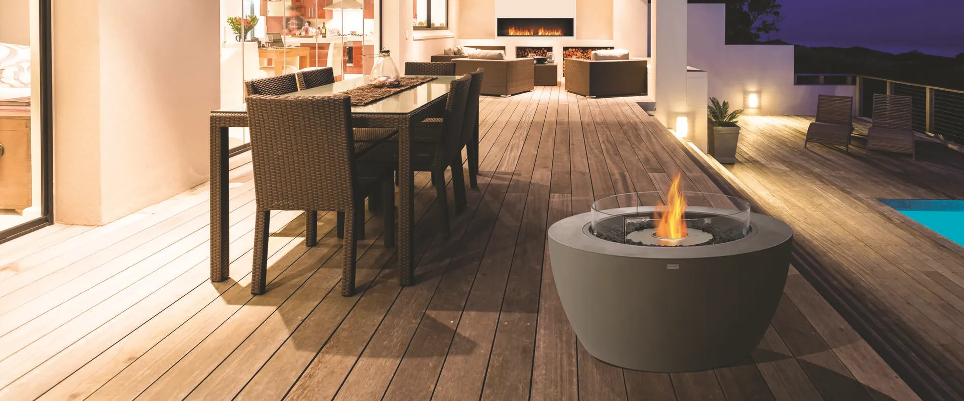 Dark EcoSmart Pod 40 bioethanol fire pit with a glass screen on a timber deck at night, beside an outdoor dining table in front of a lit modern white house