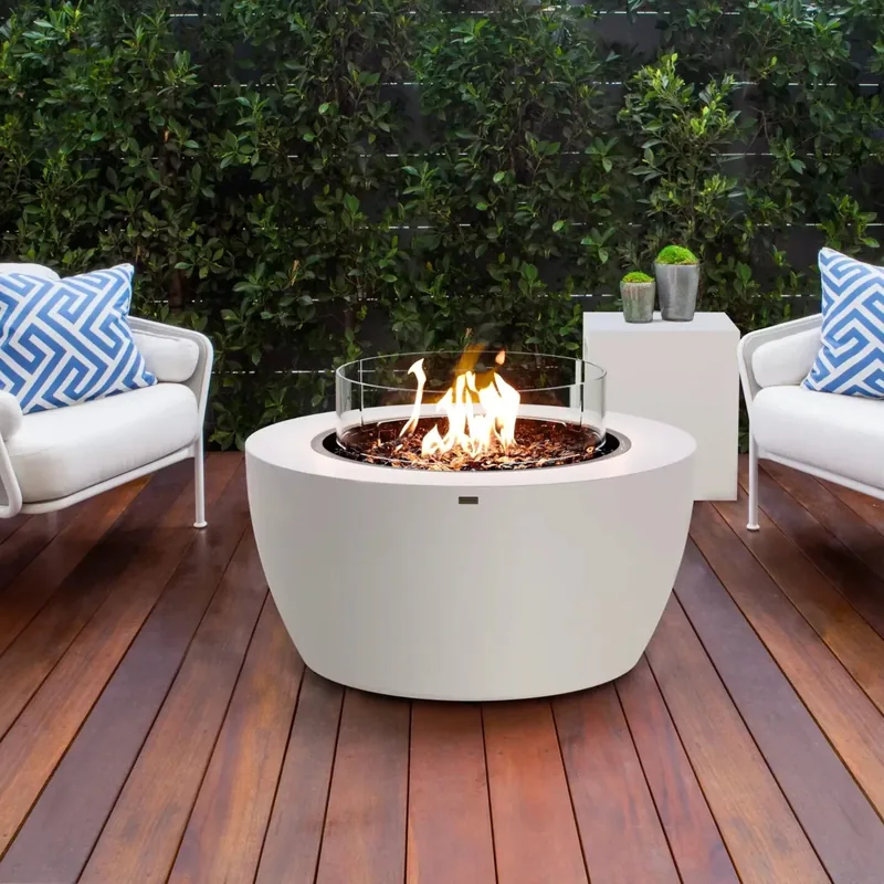 White EcoSmart Pod 40 bioethanol fire pit with a glass screen on a timber deck, between two white armchairs with blue patterned cushions in front of a green hedge