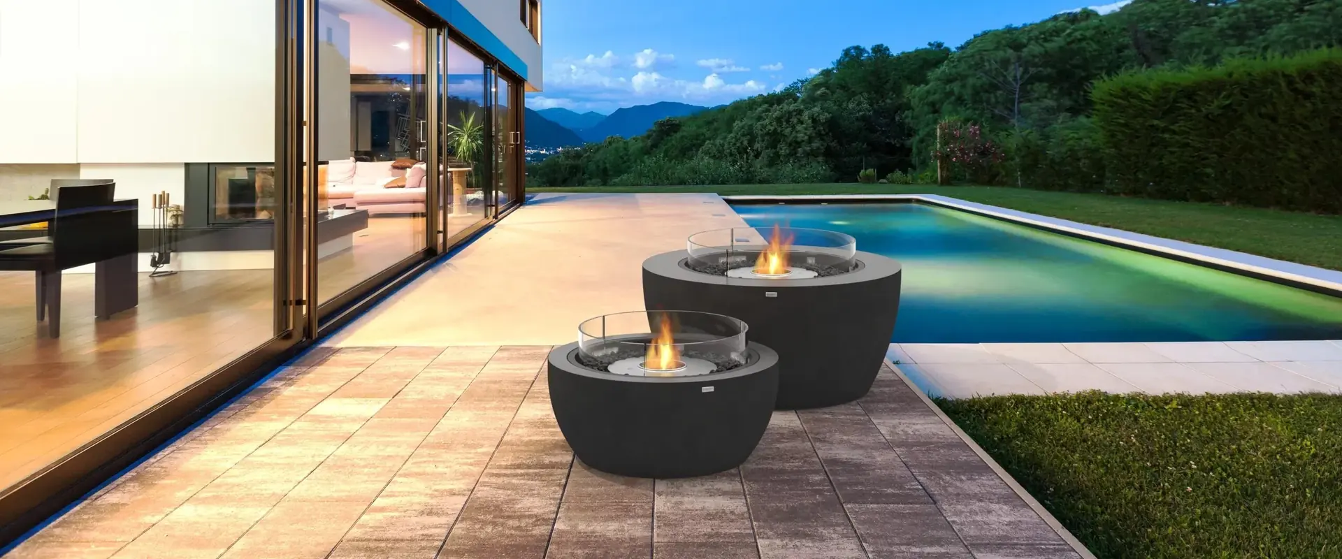 Two dark EcoSmart Pod bioethanol fire pits with glass screens on the paved terrace of a modern villa, beside a swimming pool with wooded hills beyond at dusk