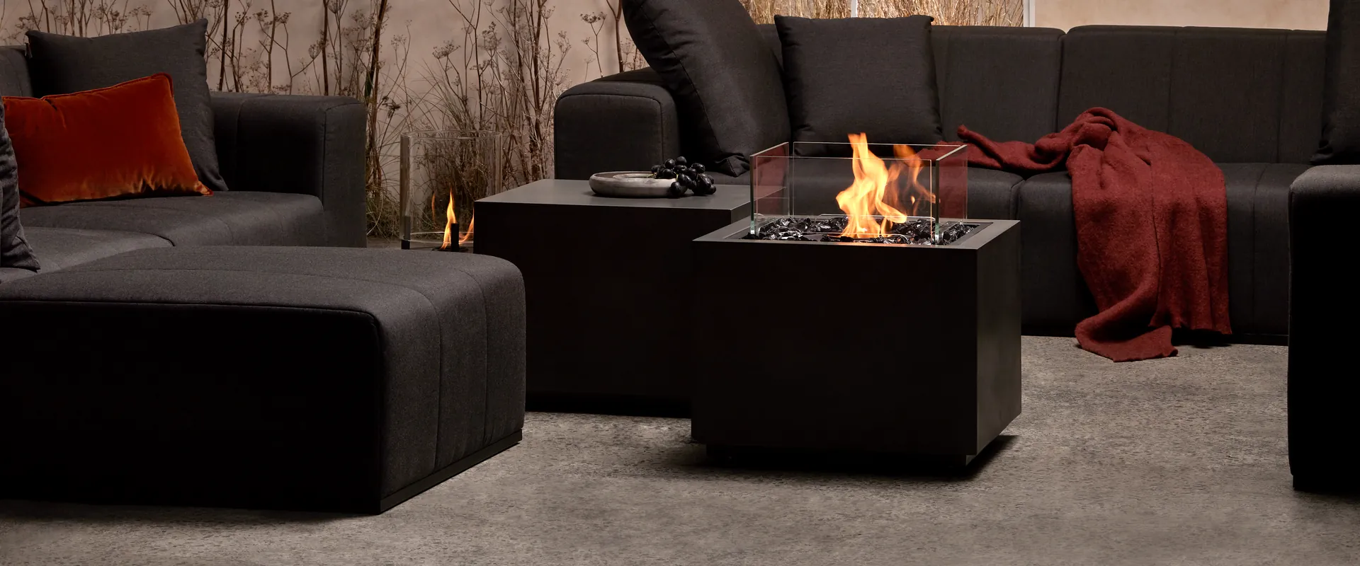 EcoSmart Sidecar 24 fire table in graphite with a glass screen burning between black modular sofas, with dried flowers behind