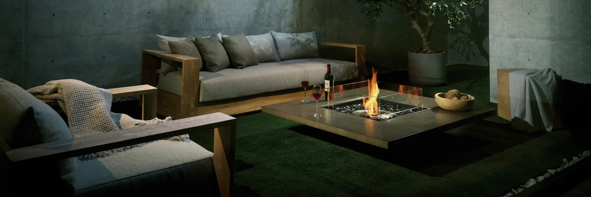 Low square EcoSmart Vertigo 40 fire table burning in front of a sofa in a room with concrete walls and a small tree, in a shaft of light