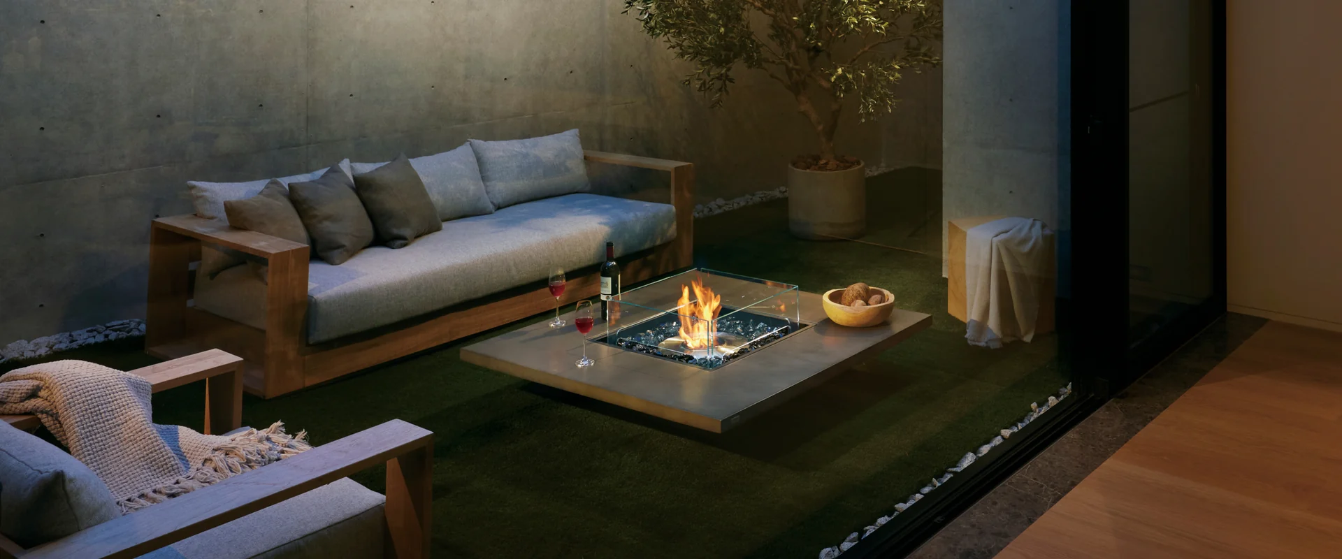 Low square EcoSmart Vertigo 50 fire table burning in front of a sofa on a patio with a small tree, seen through sliding doors