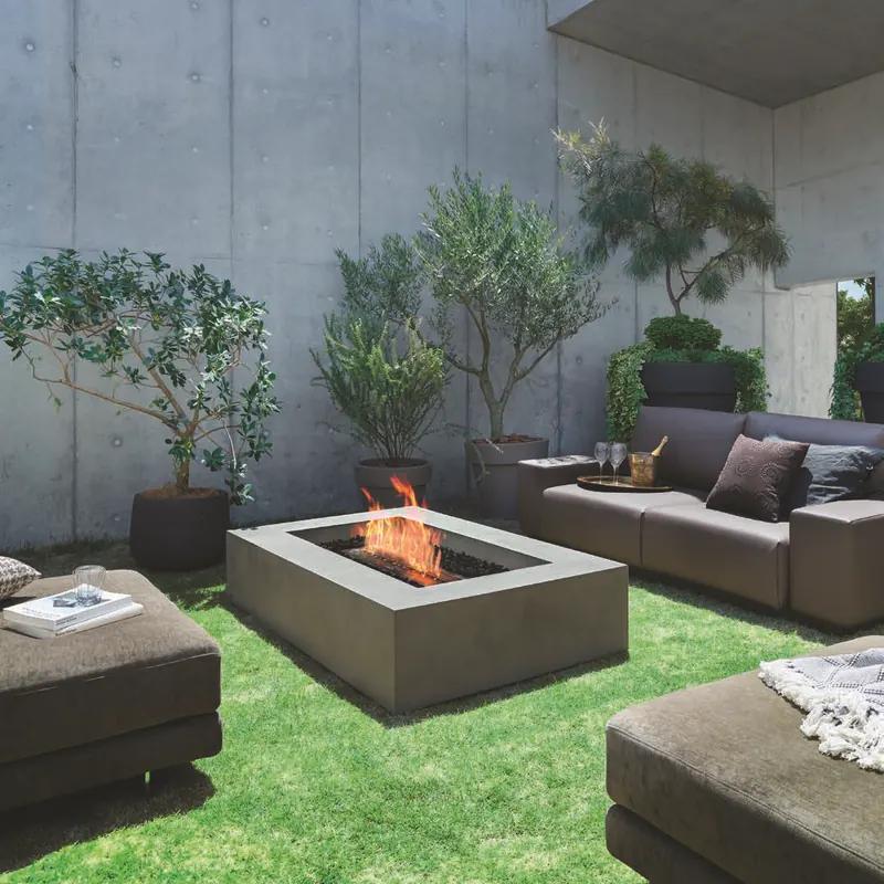 Rectangular concrete EcoSmart Wharf 65 bioethanol fire table burning on the lawn of a courtyard with concrete walls, between dark outdoor sofas and potted olive trees