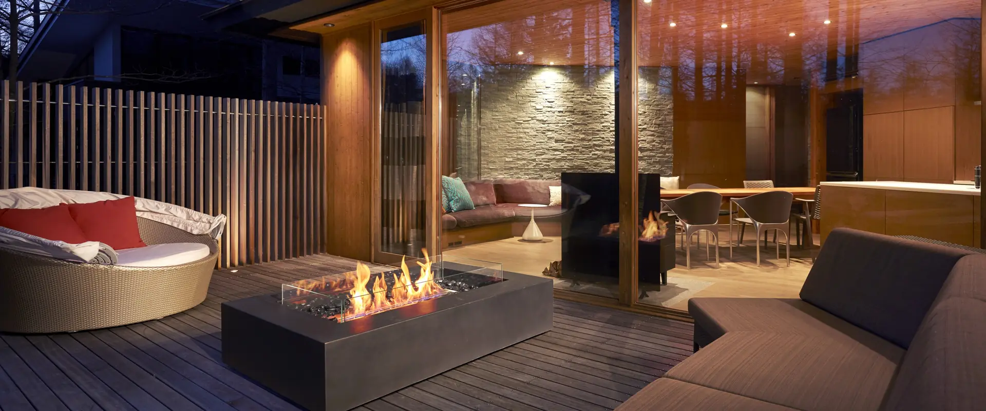 Rectangular concrete EcoSmart Wharf 65 bioethanol fire table with a line of flames on a timber deck at dusk, beside a round daybed and the lit glass doors of a timber house