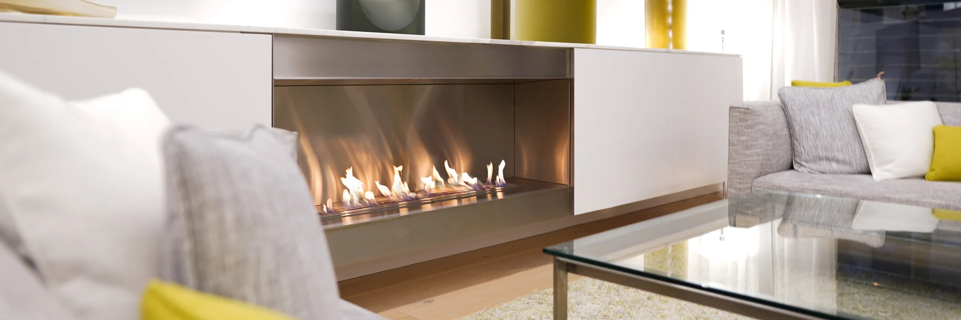 EcoSmart XL1200 bioethanol burner with a long line of flame in a white built-in fireplace, in a living room with a white sofa and yellow cushions