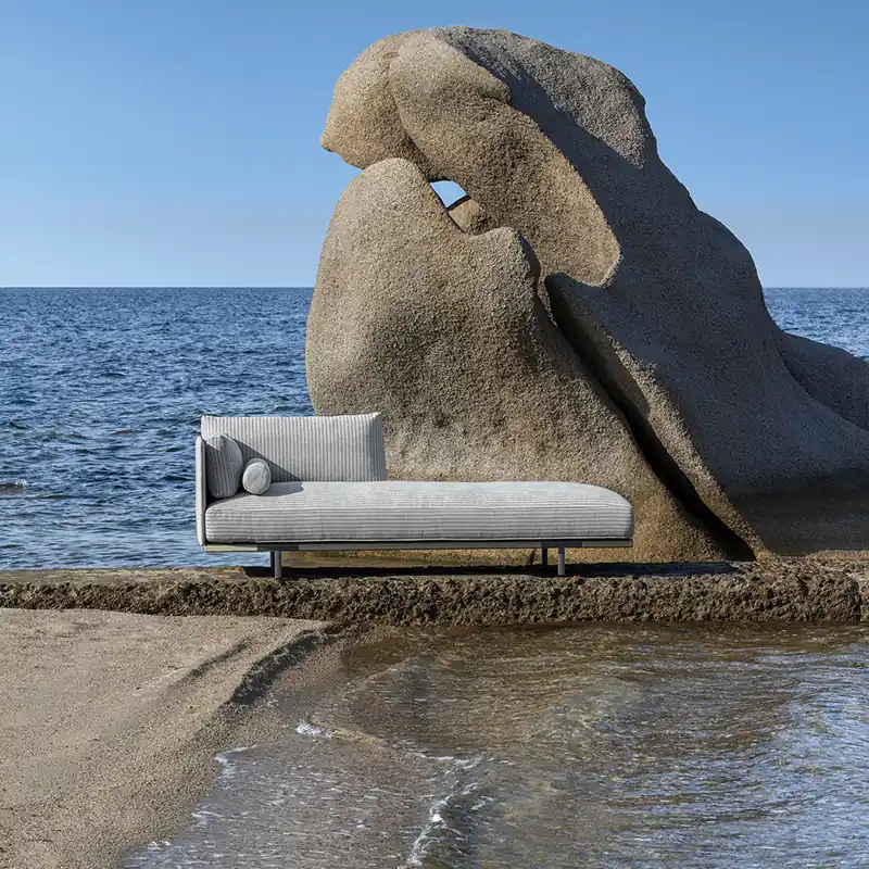 Ethimo Baia sofa standing on the sand at the water's edge beside a wind-carved granite rock