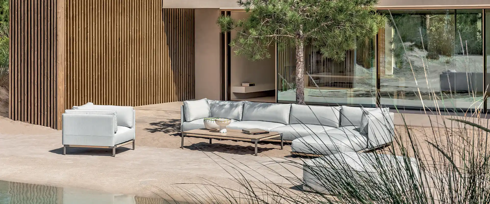 Ethimo Baia curved modular sofa and armchair in pale fabric on the sand-colored terrace of a modern villa with pines