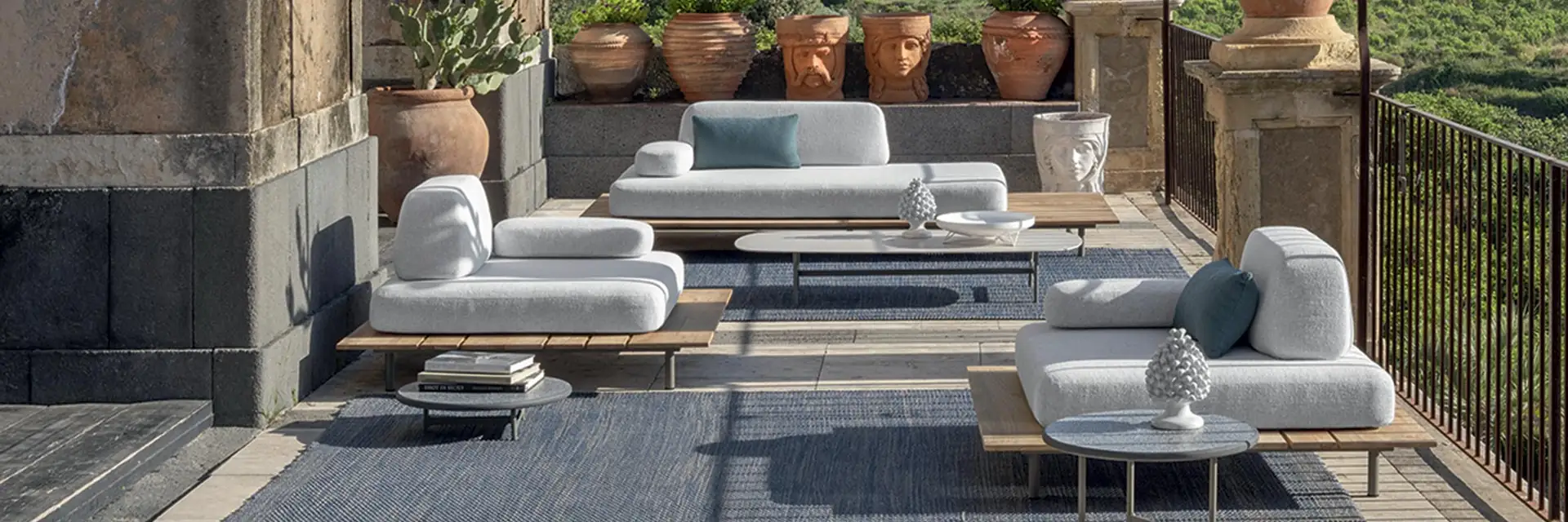 Ethimo Calipso modular lounge with pale cushions on a terrace with terracotta pots and an iron railing, hills behind