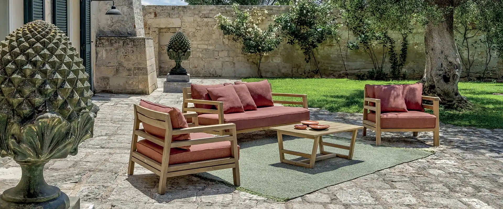 Ethimo Costes teak sofa and armchairs with red cushions on a stone-paved courtyard with a lawn and old stone walls